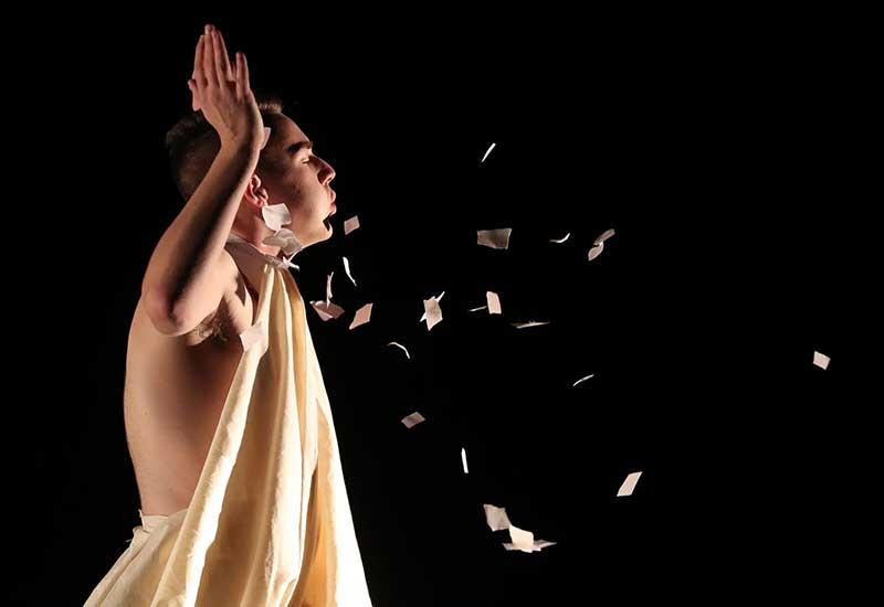 a performer wearing a toga blows scraps of paper across the air during a dance performance